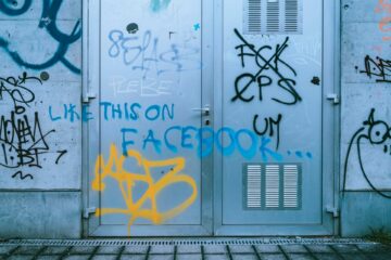 Graffiti "Like this on Facebook" na zniszczonych metalowych drzwiach, symbolizujące minioną erę "starego Facebooka" i jego wpływ na kulturę.