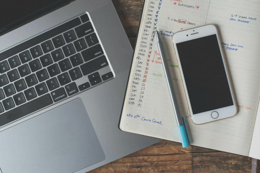 Widok z góry na biurko z otwartym laptopem MacBook Pro, obok leży smartfon iPhone, długopis oraz otwarty kalendarz/notes z ręcznymi zapiskami i harmonogramem. Zdjęcie symbolizuje proces planowania, zarządzania czasem i tworzenia treści cyfrowych.